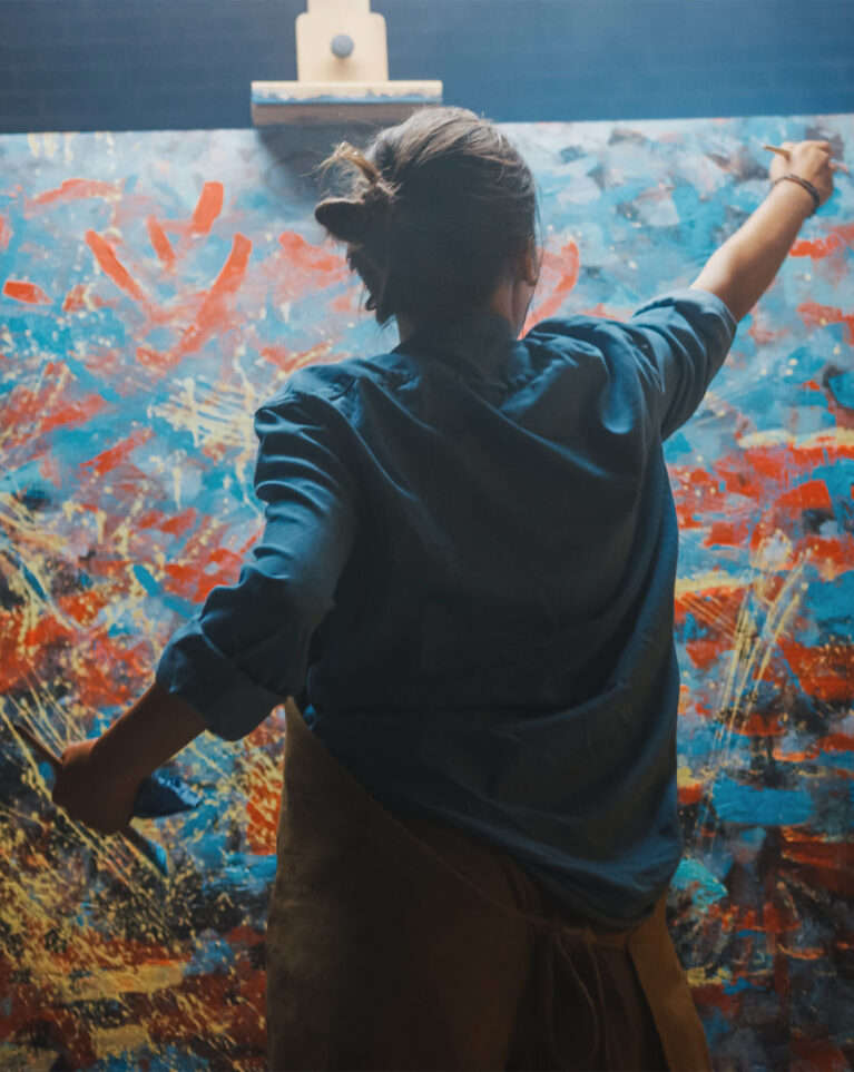 Talented Female Artist Works on Abstract Oil Painting, Using Paint Brush She Creates Modern Masterpiece. Dark and Messy Creative Studio where Large Canvas Stands on Easel Illuminated.