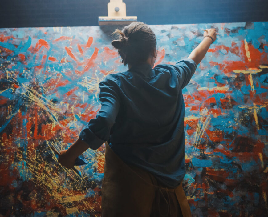 Talented Female Artist Works on Abstract Oil Painting, Using Paint Brush She Creates Modern Masterpiece. Dark and Messy Creative Studio where Large Canvas Stands on Easel Illuminated.